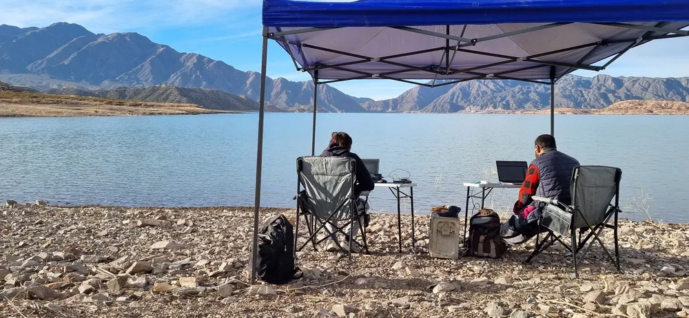 Dos personas trabajando con sus notebooks bajo un gazebo, frente a un lago y la cordillera